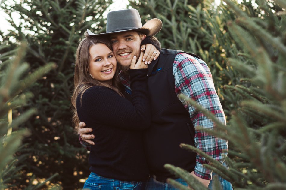 A couple embracing outdoors in a wooded area with evergreen trees. The man is wearing a gray cowboy hat and plaid shirt, and the woman is wearing a black sweater. Both are smiling.