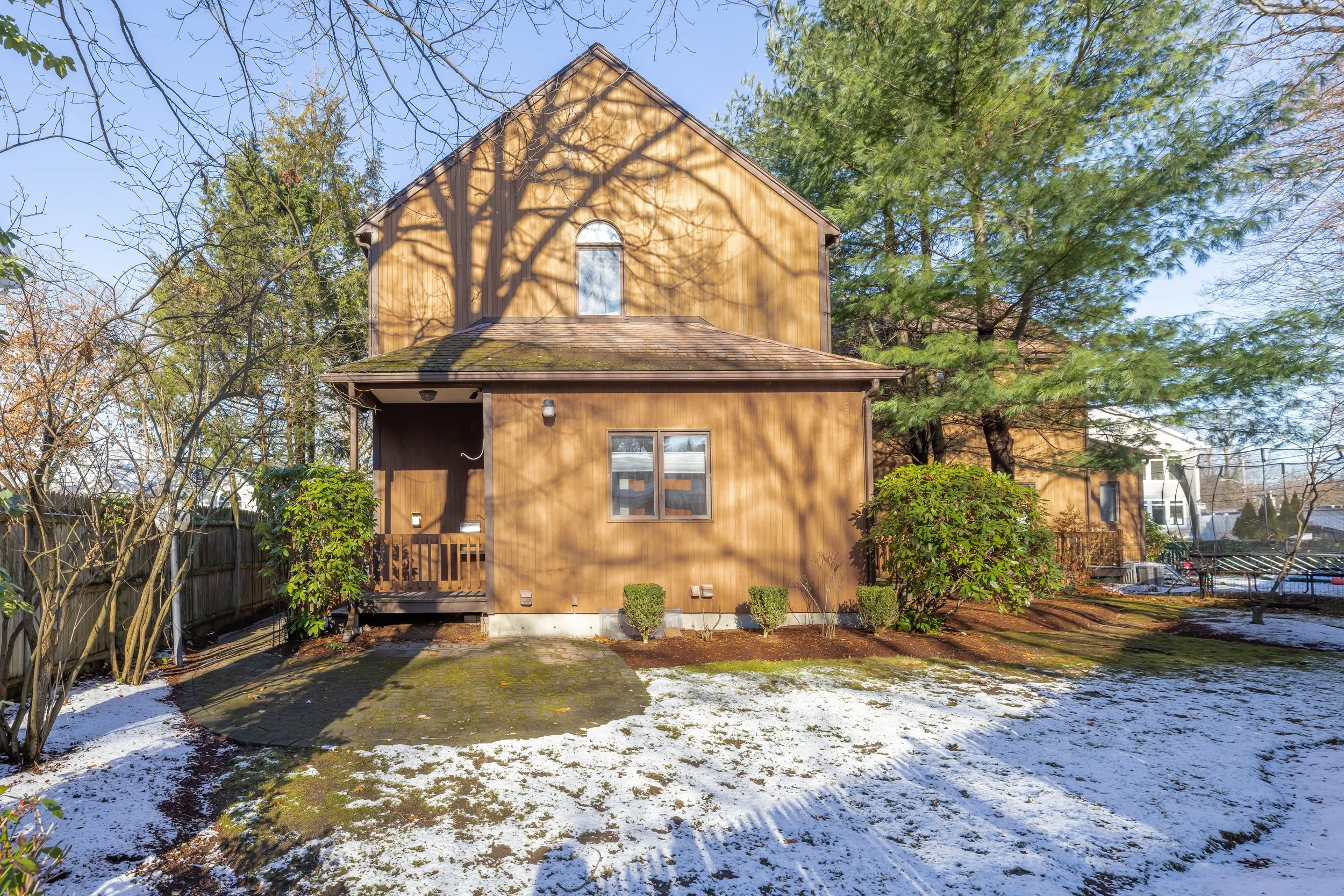 Front view of a two-story wooden house with a small porch, surrounded by trees and patches of snow on the ground.