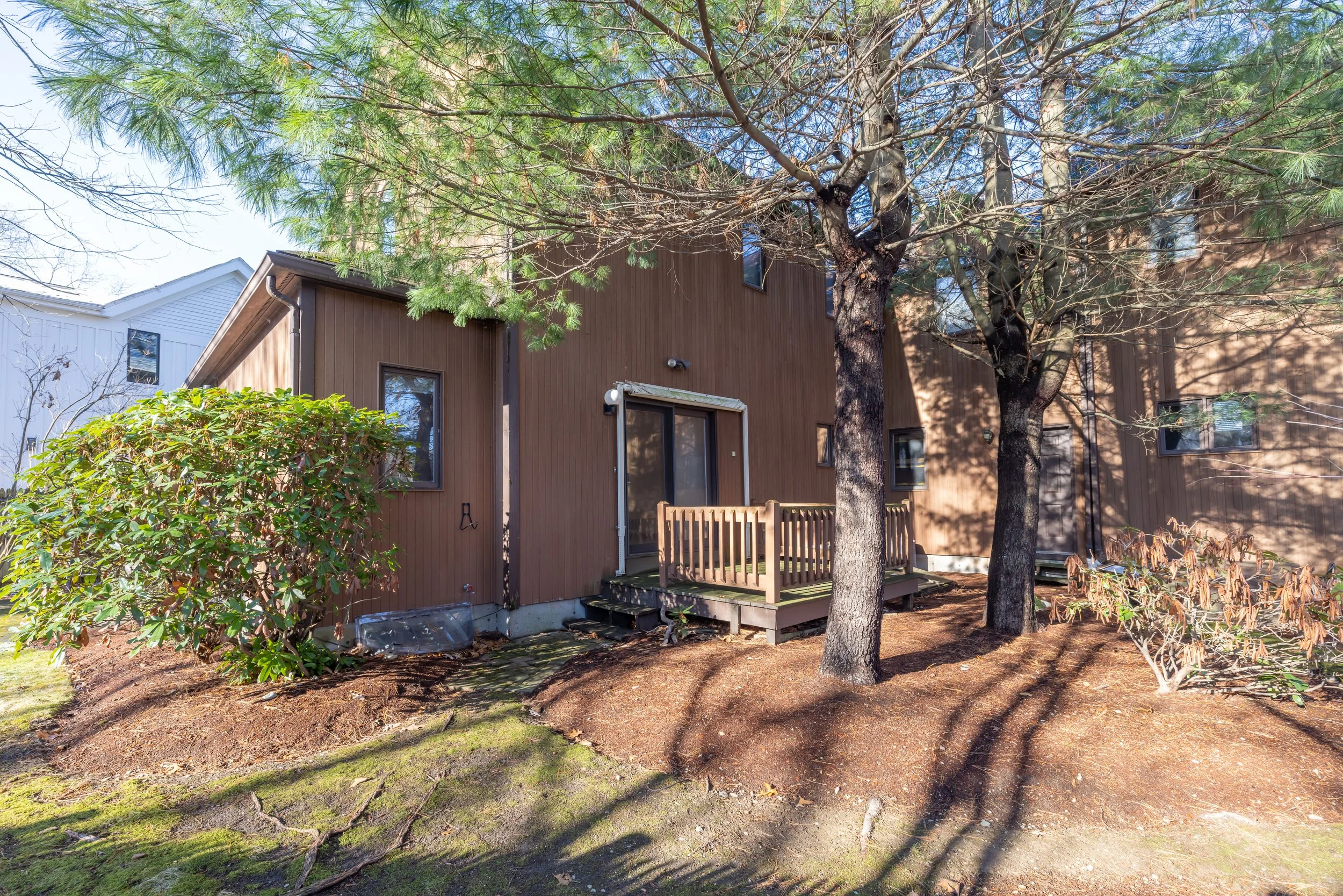 Back of a brown house with a small wooden deck, surrounded by trees and shrubs, with shadows cast on the ground.