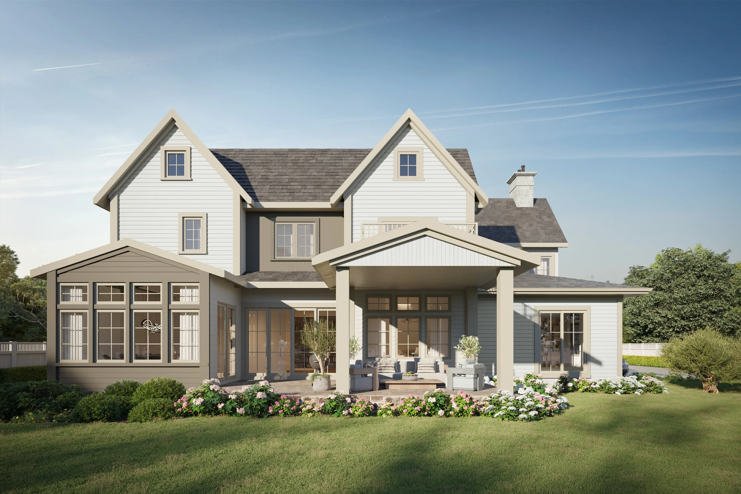 A large, modern two-story house with gray siding and a gabled roof, featuring a covered porch with outdoor seating, surrounded by a green lawn and blooming flowers.