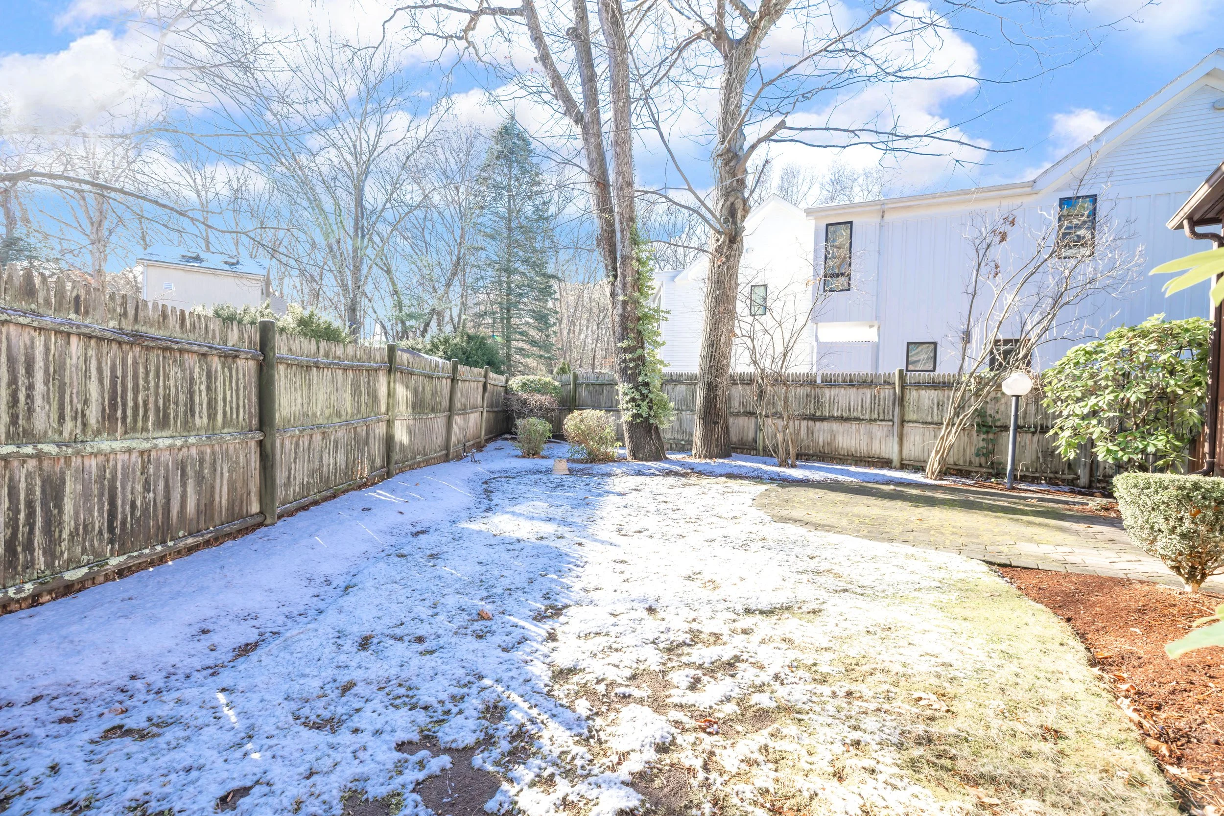 A backyard with a wooden fence, snow on the ground, trees without leaves, and a white house in the background under a partly cloudy sky.