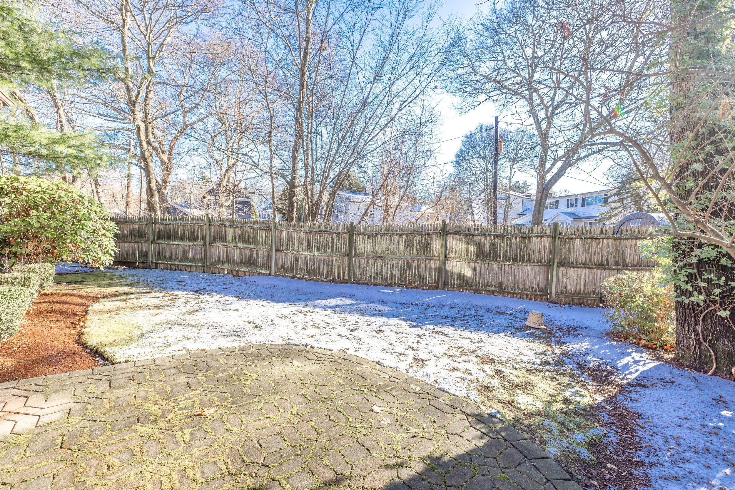 Backyard view with a brick patio, a snow-covered lawn, leafless trees, and a wooden fence, under a clear blue sky.