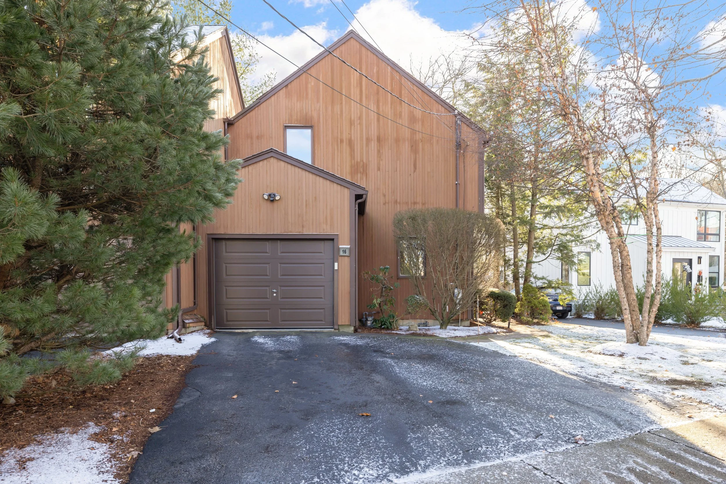 A modern, two-story house with a wooden exterior, a brown garage door, and a driveway with patches of snow. There are trees and shrubs around the house, with some snow on the ground and a partly cloudy sky above.
