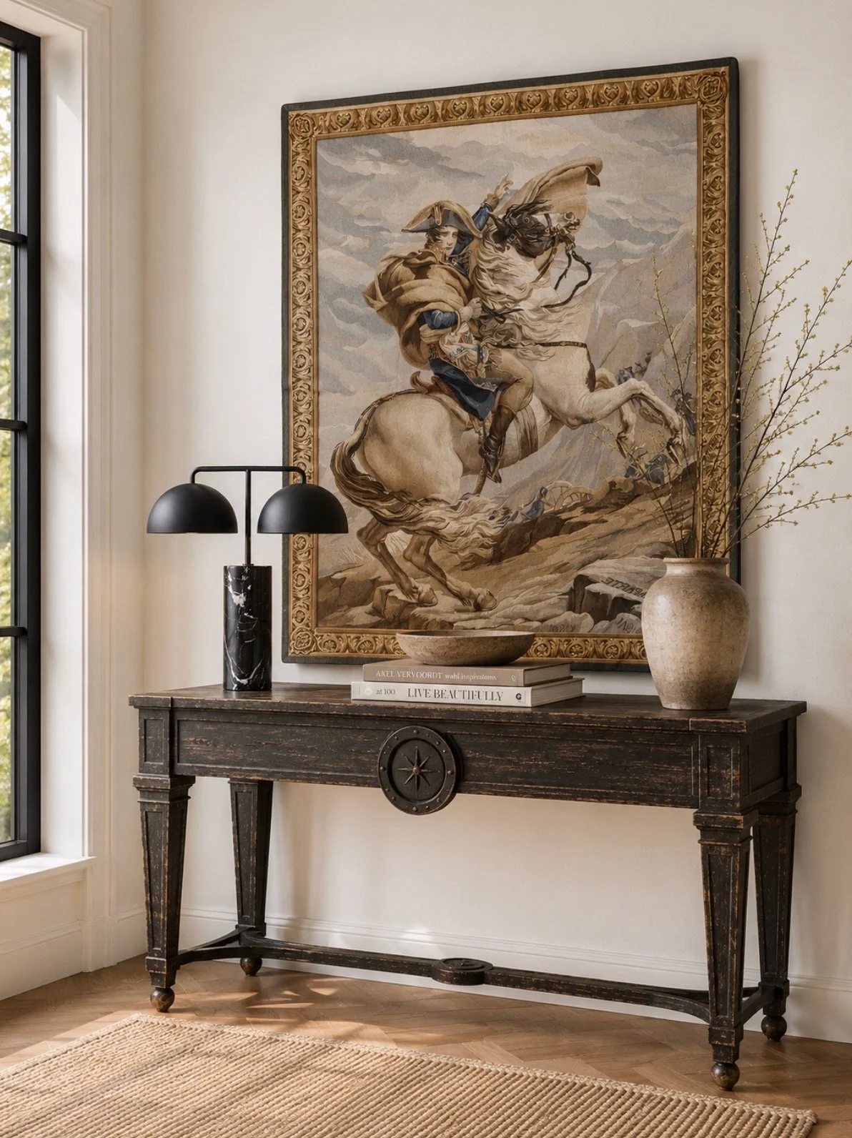 A living room entryway with a large framed tapestry of Napoleon on horseback, a black and white marble table lamp, a beige vase with twigs, and stacked books on a dark wooden console table near a window with a view of trees.