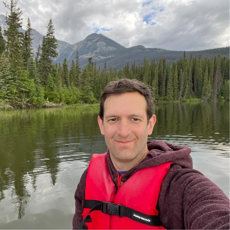 A man taking a selfie on a calm lake with pine trees and mountains in the background during the daytime, wearing a red life jacket and a brown hoodie.