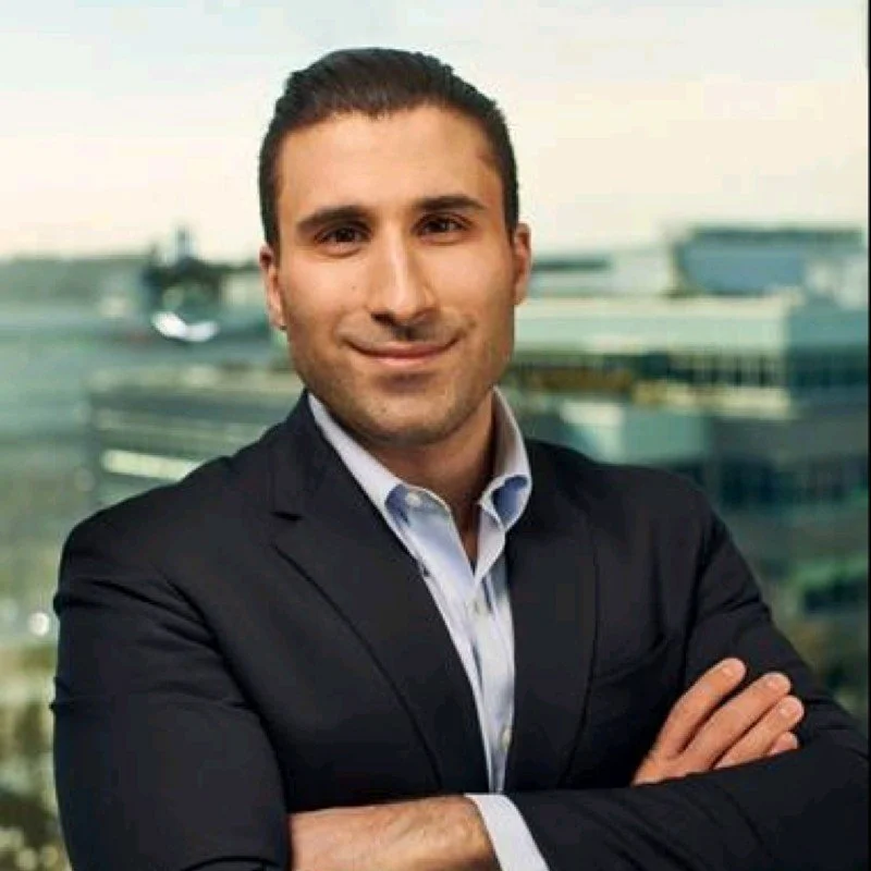 A man in a business suit with crossed arms standing in front of a window with a cityscape background.