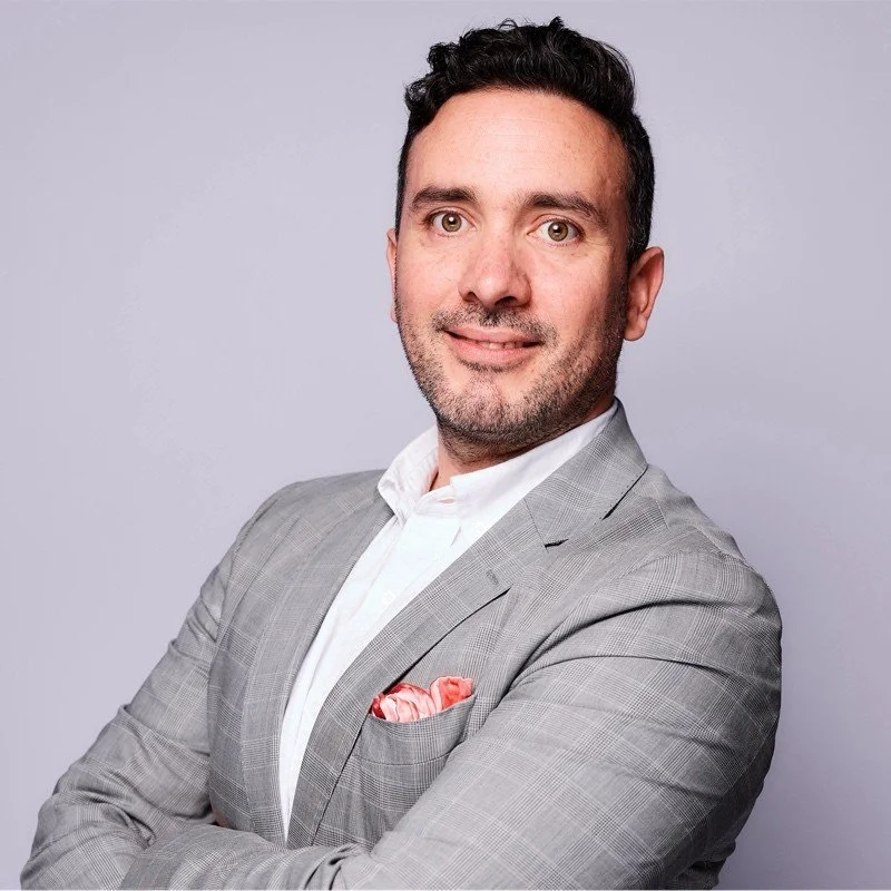 A man with dark hair and light skin, wearing a gray suit with a white shirt and a pink pocket square, stands against a plain gray background.