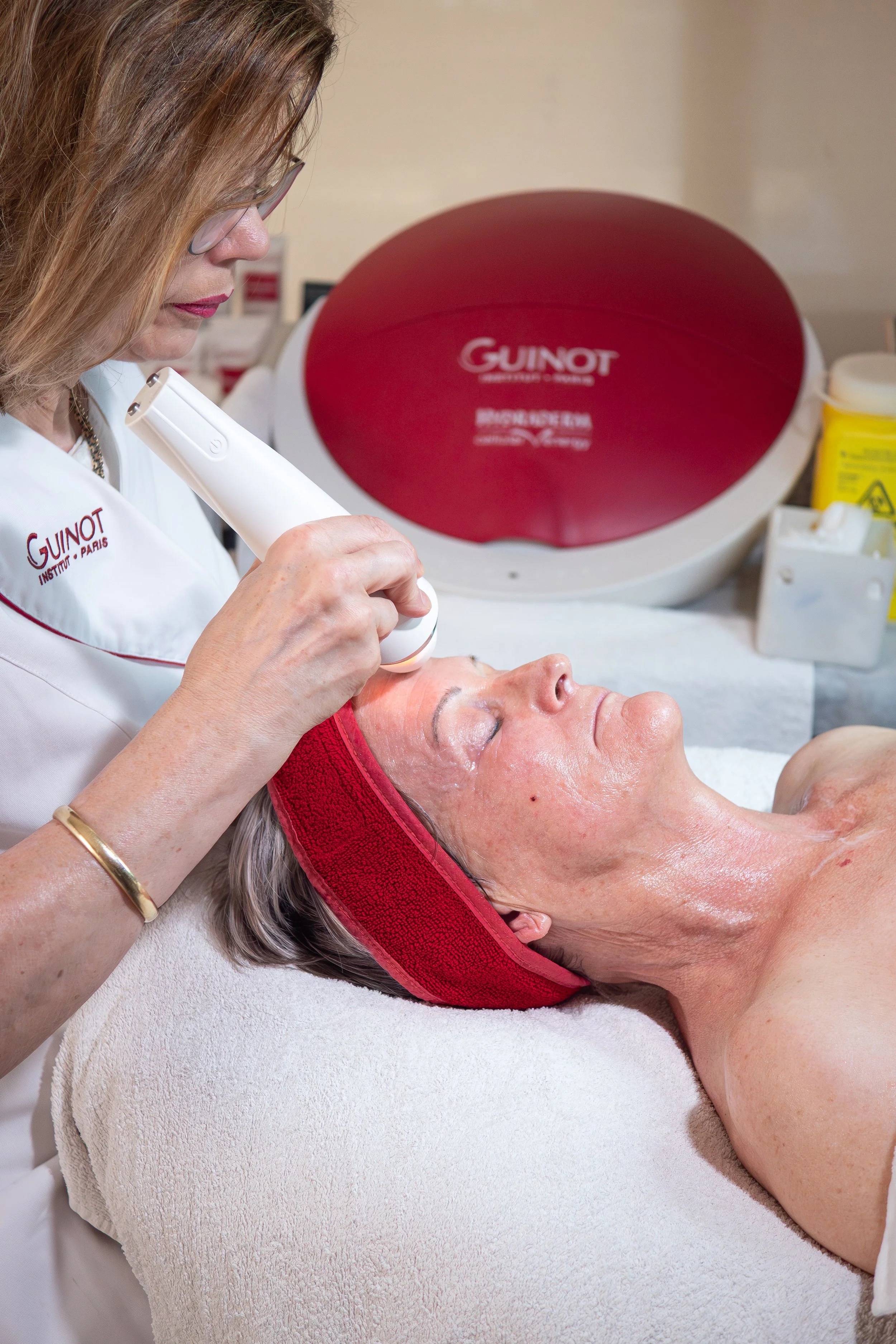 A woman receives a facial treatment from a cosmetologist using a handheld device in a spa or clinic setting.