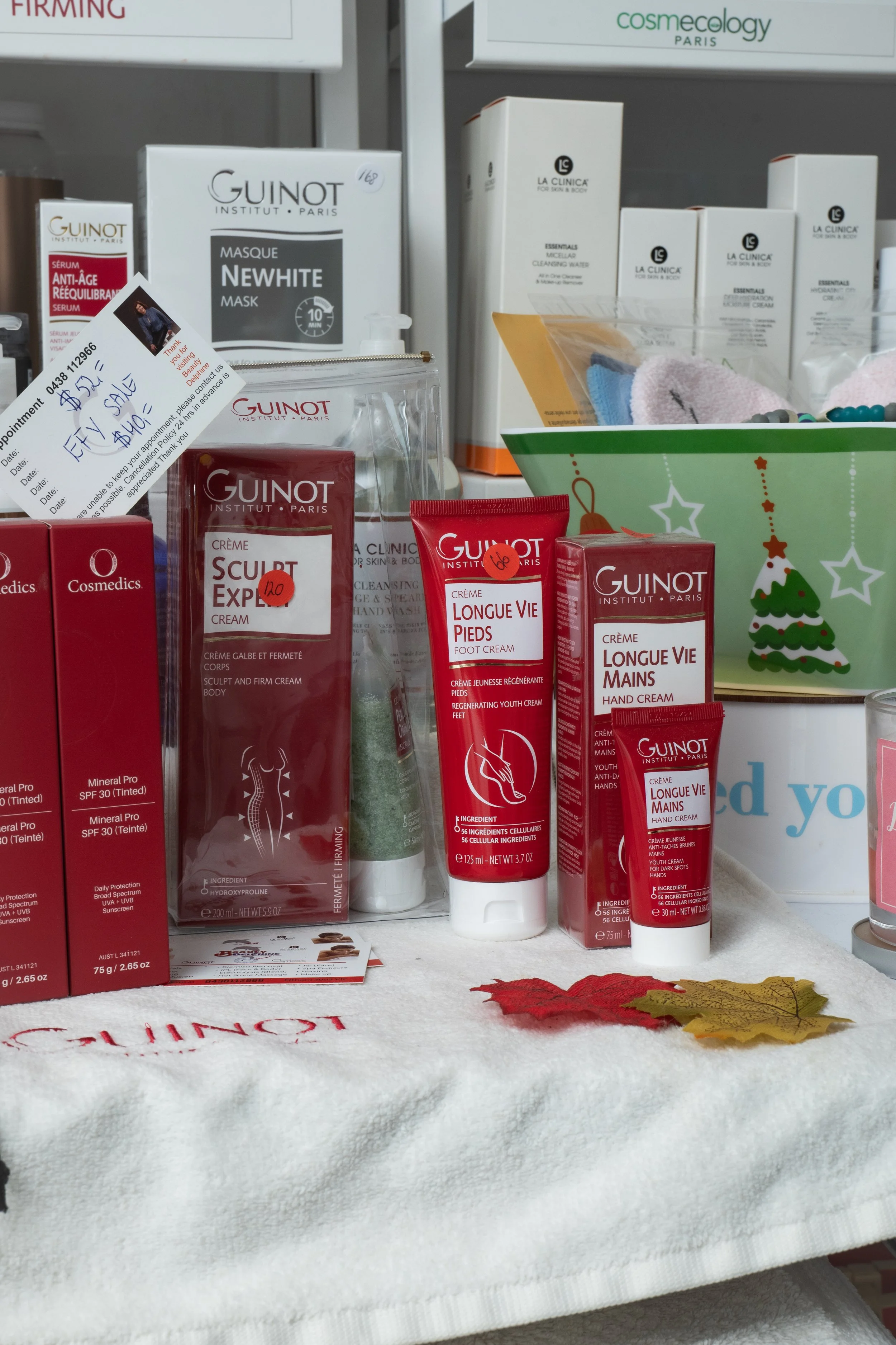Display of Guinot skincare products, including creams and foot and hand treatments, placed on a white cloth with autumn leaves and a holiday-themed gift bag in the background.