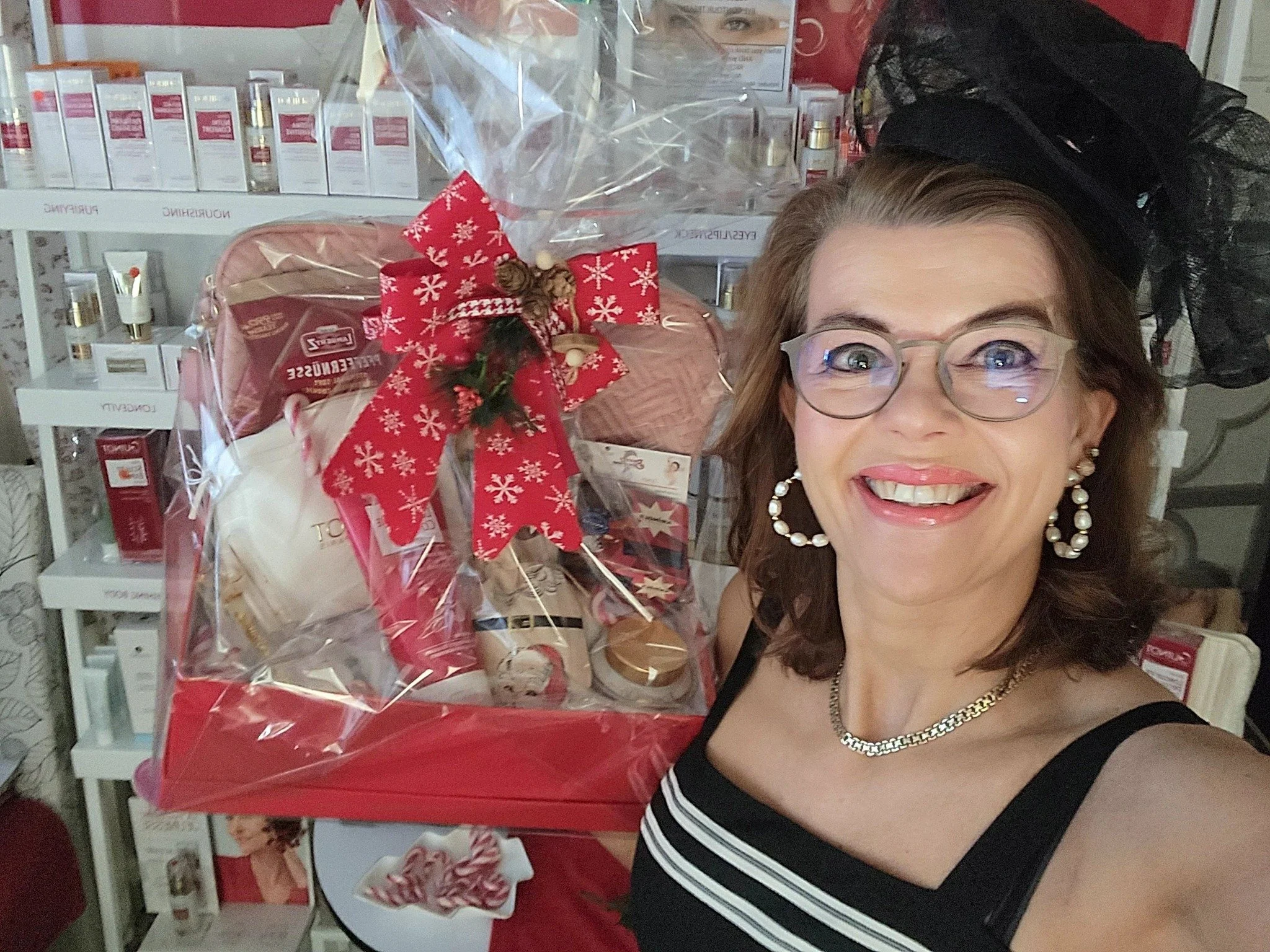 A woman with glasses, hoop earrings, and a black and white striped dress smiling for a selfie next to a large Christmas gift basket wrapped in clear cellophane and decorated with a red bow, in a store with shelves of skincare products in the background.