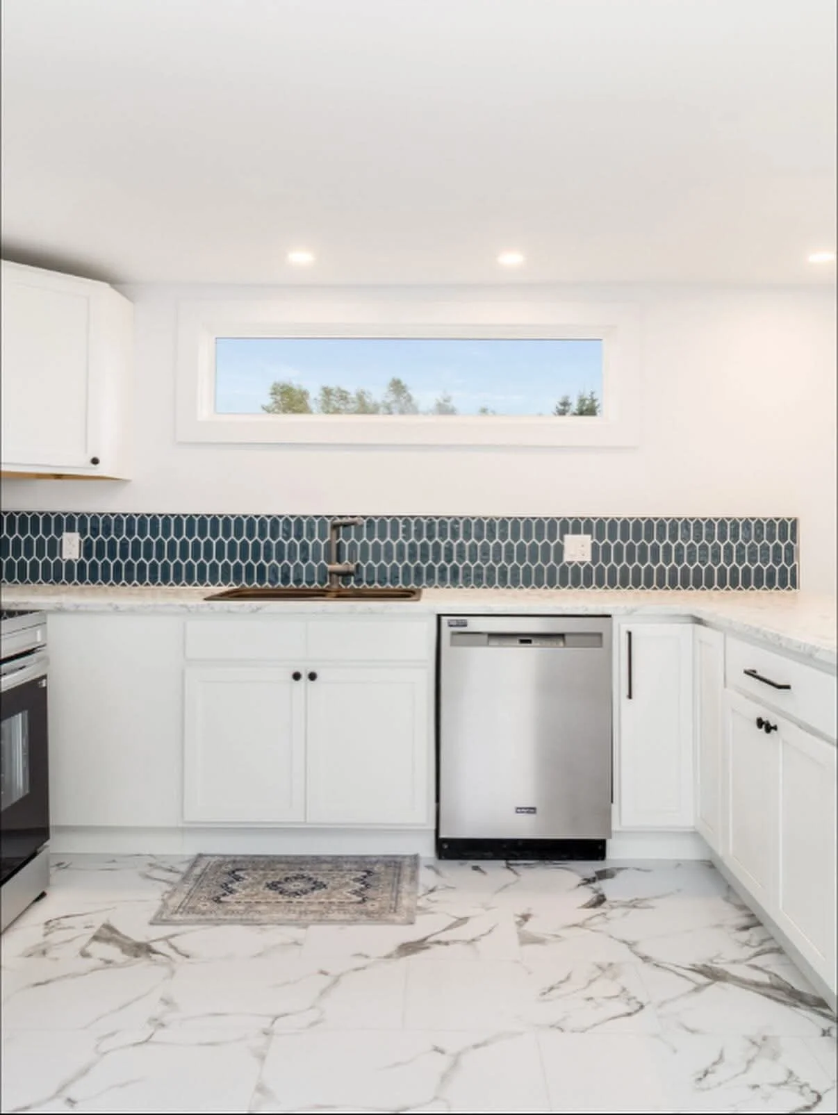 Modern white kitchen with marble countertops, blue hexagonal backsplash, and stainless steel appliances, including dishwasher and oven, with a small woven rug on marble floor and a horizontal window above the sink.