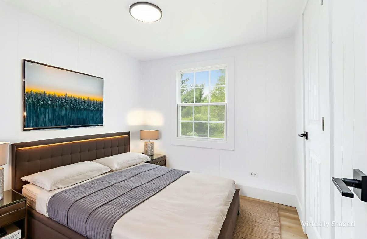 A minimally decorated bedroom with a bed, two bedside tables with lamps, a window showing trees outside, a wall-mounted television, and a door, with white walls and a beige rug.