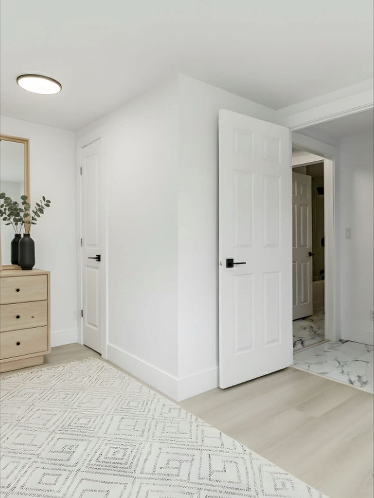 Interior view of a white room with a corner, showing two white doors, a beige dresser with black vases and green foliage, a mirror, a patterned white rug, and white flooring with a marble transition area.