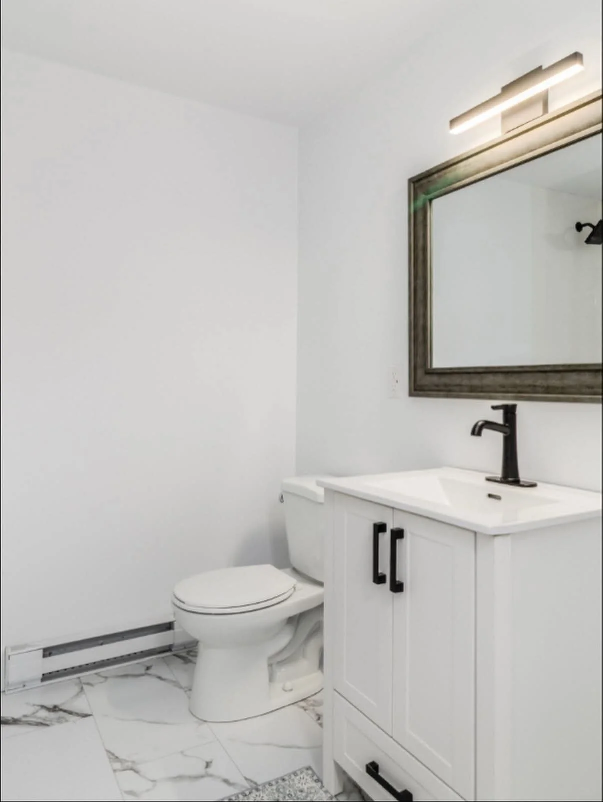 A bathroom with white walls and marble tile flooring, featuring a toilet, a white vanity with a black faucet, a large wooden framed mirror, and a modern light fixture above the mirror.