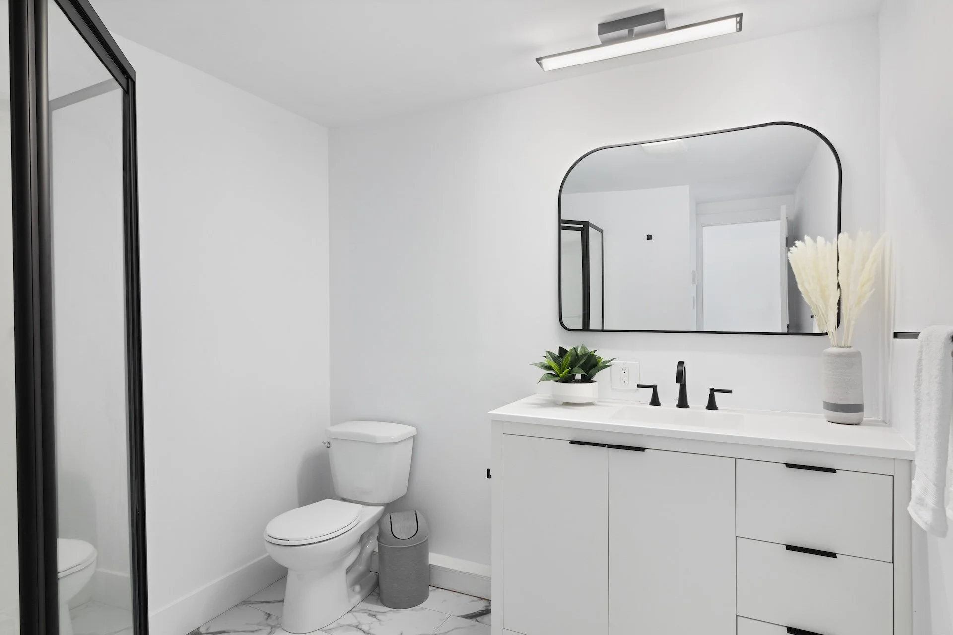 A minimalist bathroom with white walls, a white vanity with black handles, a large black-framed mirror, a potted plant, and a vase with white dried flowers. There is a toilet and a glass shower enclosure with black framing. The floor has white marble