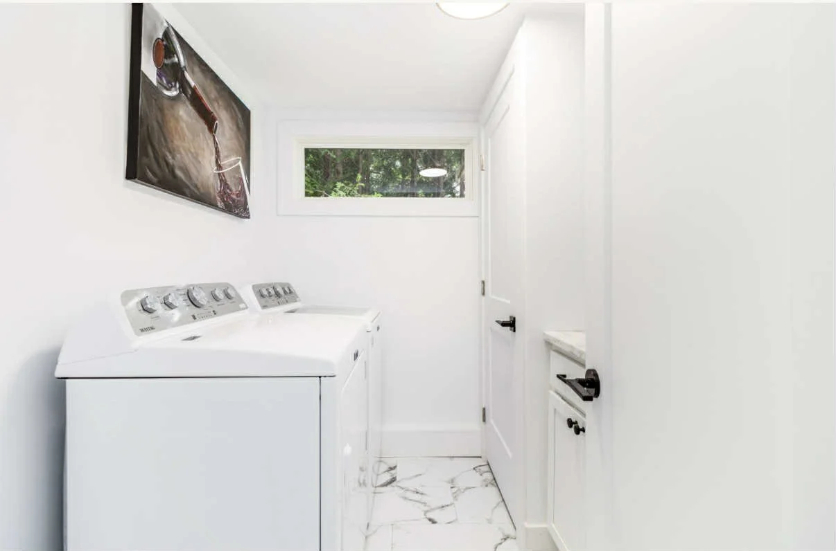 A small laundry room with white walls, a white top-loading washing machine, a white cabinet with a marble countertop, a small window, and a wall-mounted flat-screen TV with a wine glass and bucket of wine on the screen.