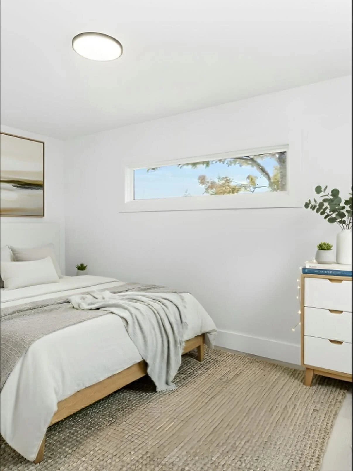 A minimalistic bedroom with white walls and bedding, a light wooden bed frame, a beige woven rug, and a white dresser decorated with potted plants. There is a horizontal window with a view of trees outside.