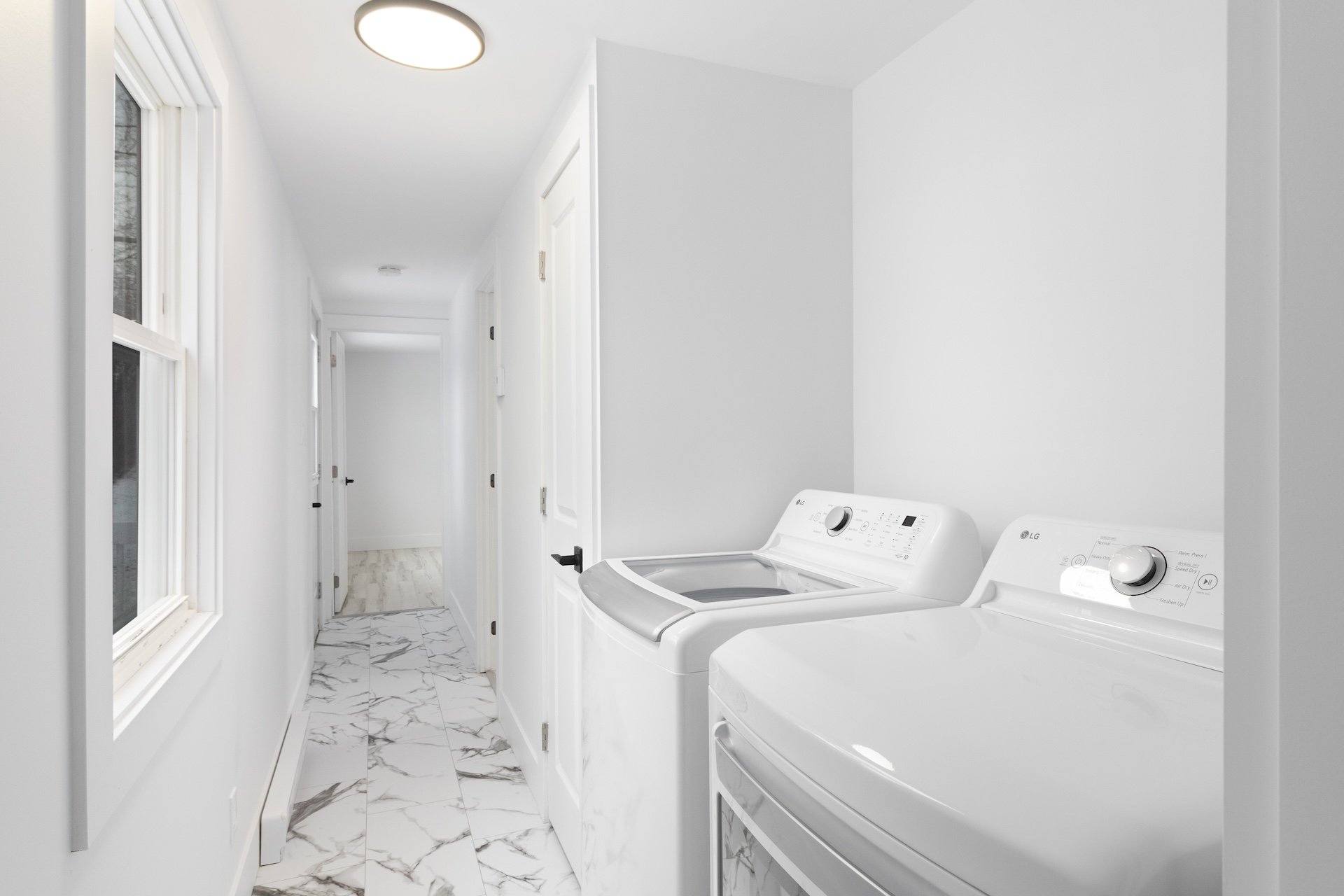 Laundry room with white walls, marble tile floor, window on the left, and a washing machine and dryer on the right.