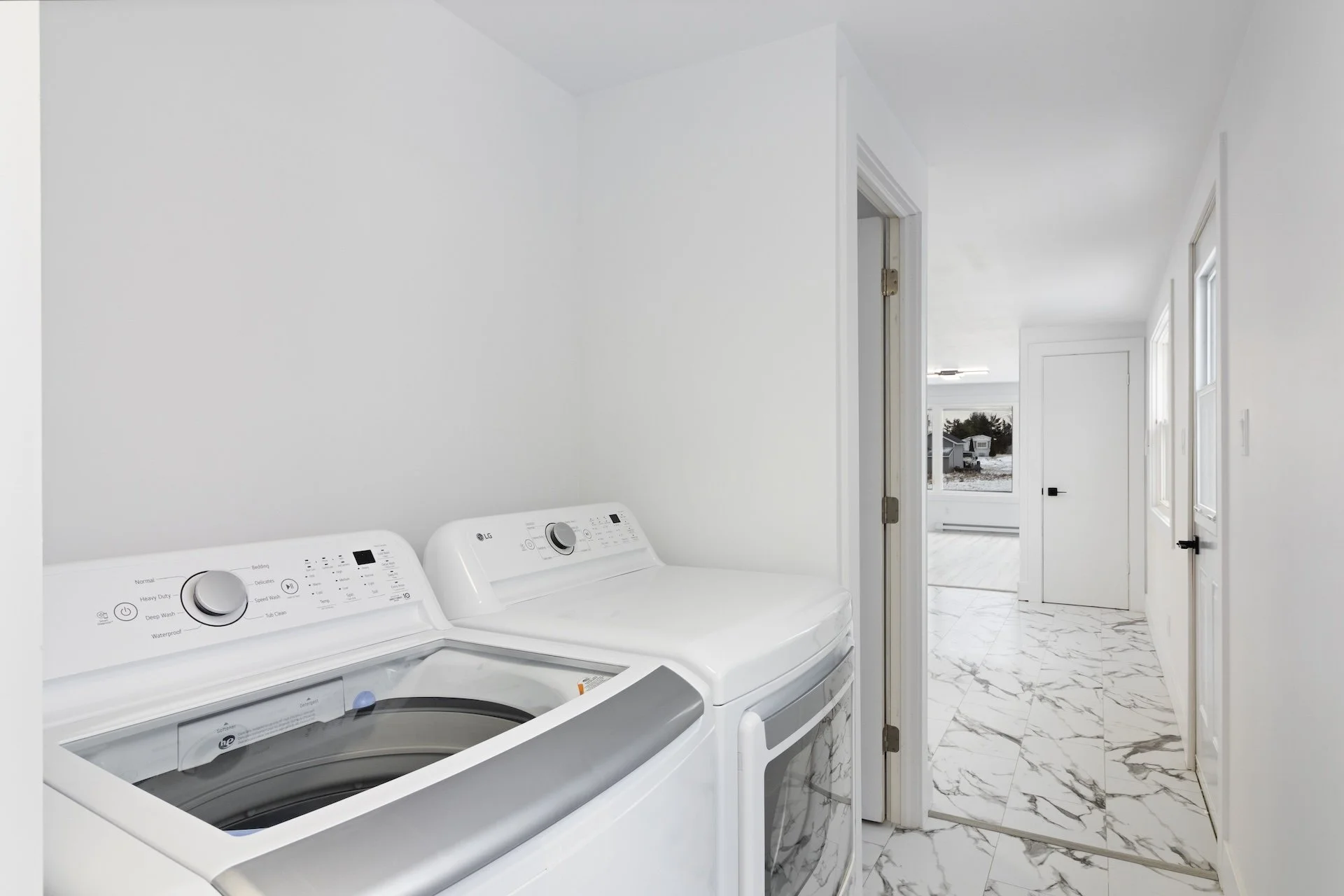 Laundry room with a washing machine and dryer against a white wall, white marble floor, open door leading to another room with windows showing an outdoor scene.