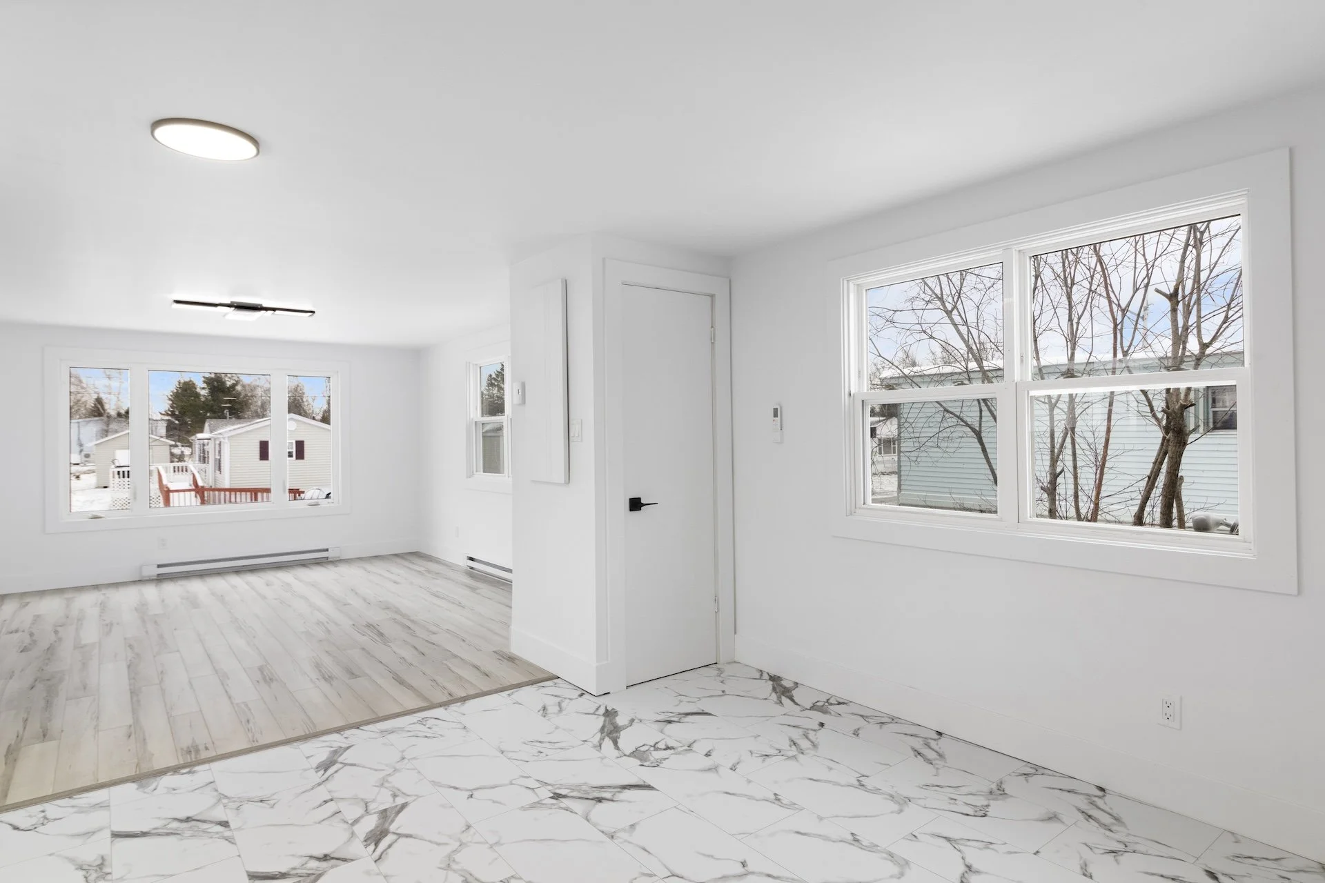Empty bright living room with white walls and large windows showing snow outside, wood and marble flooring, and minimal ceiling lights.