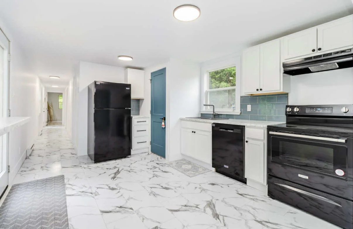 Modern kitchen with white cabinets, black appliances, and a blue door. Marble flooring, gray tile backsplash, and a window above the sink.