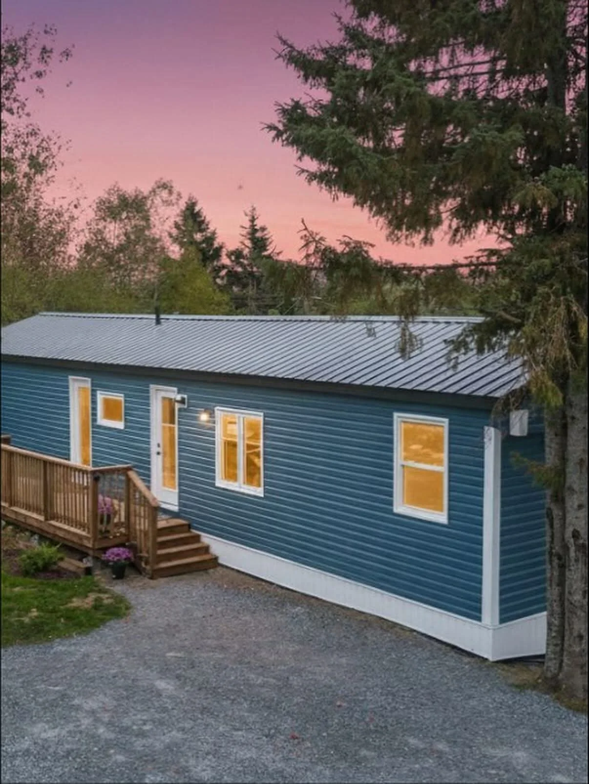 A blue house with white trim, a metal roof, and a wooden porch with stairs, illuminated from inside, set against a pink and purple sunset sky with tall trees surrounding it.