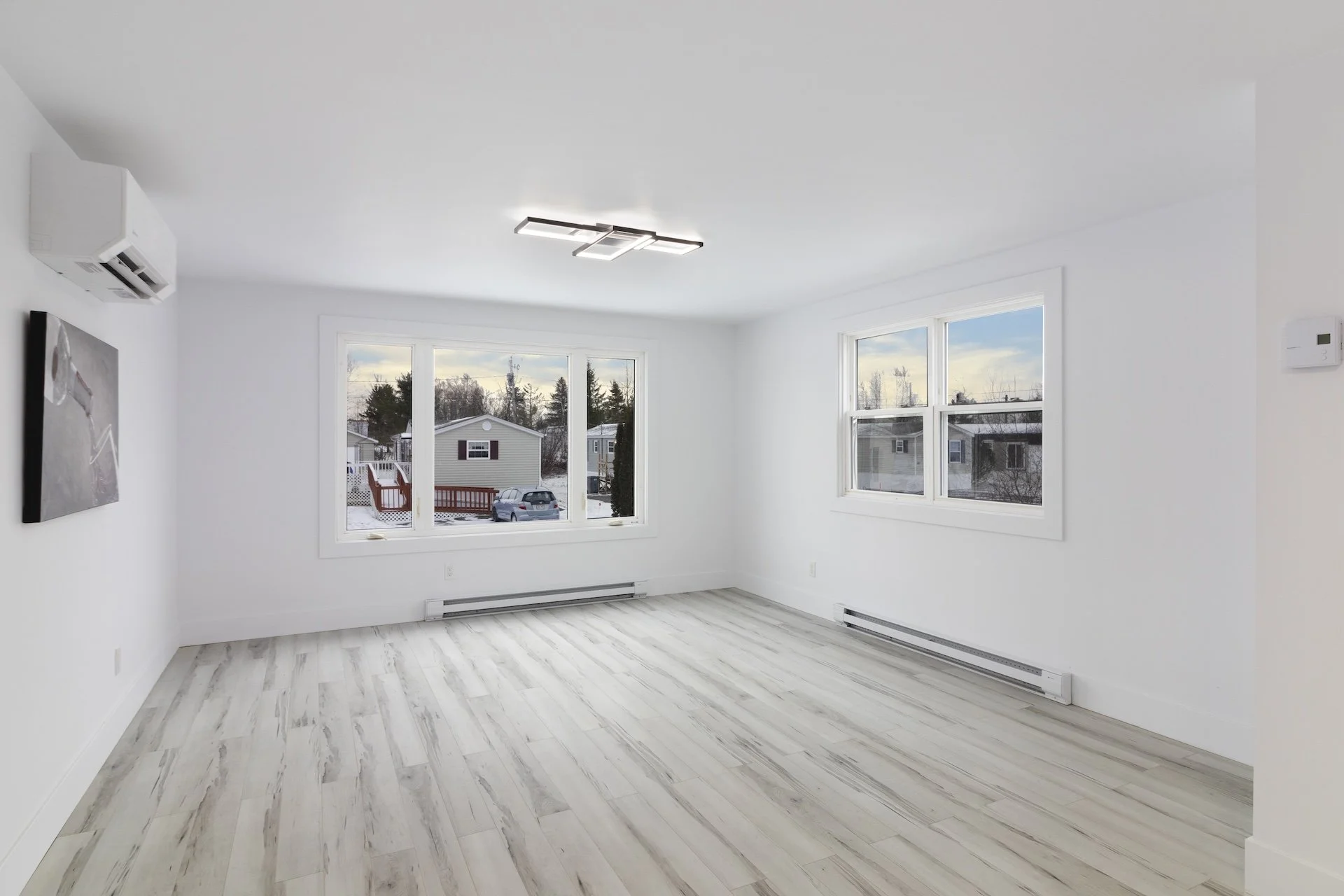 Empty living room with white walls, light-colored wood floor, two large windows showing a snowy outside scene, modern ceiling light fixture, wall-mounted air conditioning unit, and heating baseboards.