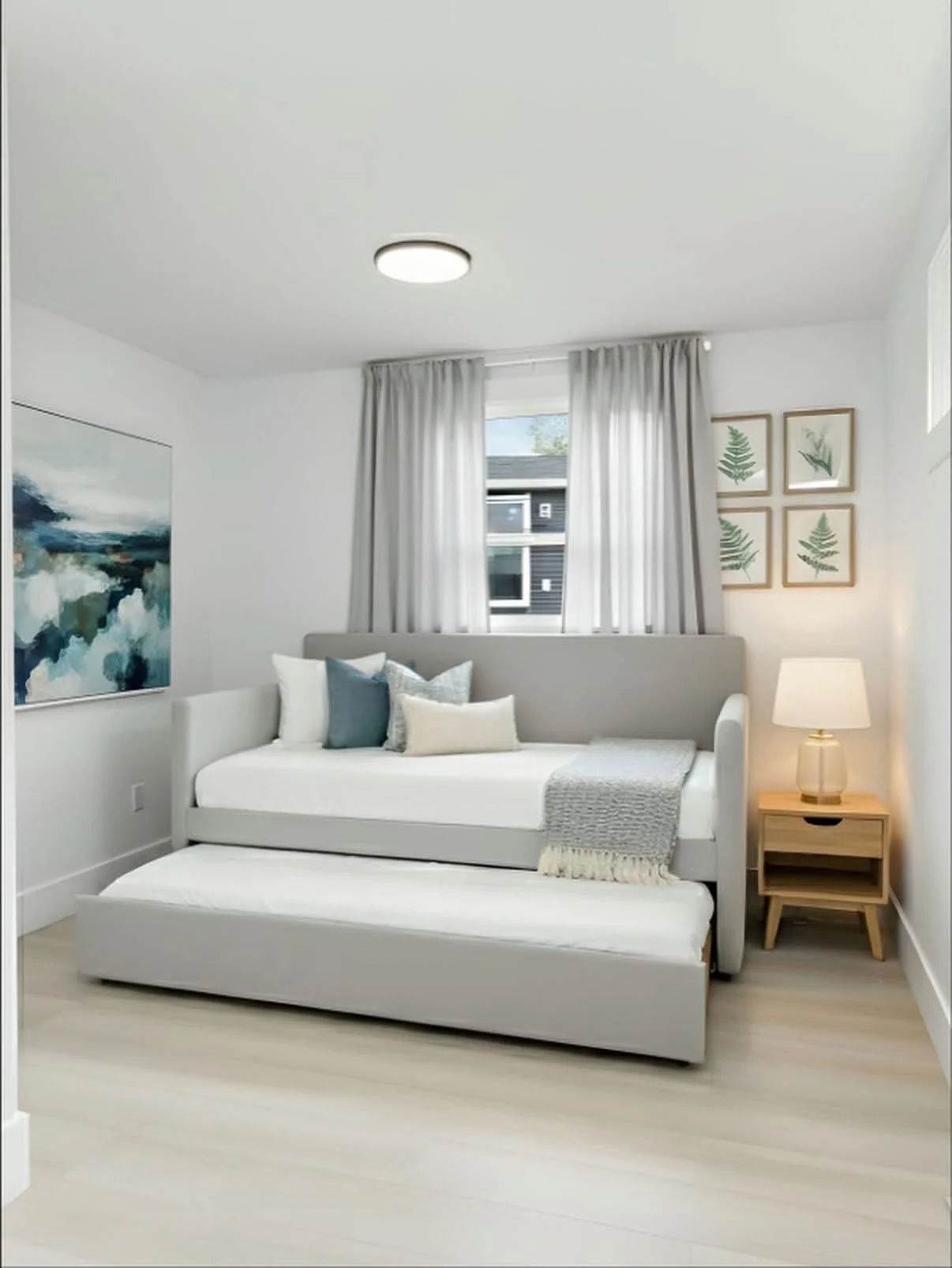 A bright modern bedroom with white walls, light wood flooring, a bed with white and gray bedding, a nightstand with a lamp, wall art of green leaves, and a large window with white curtains.