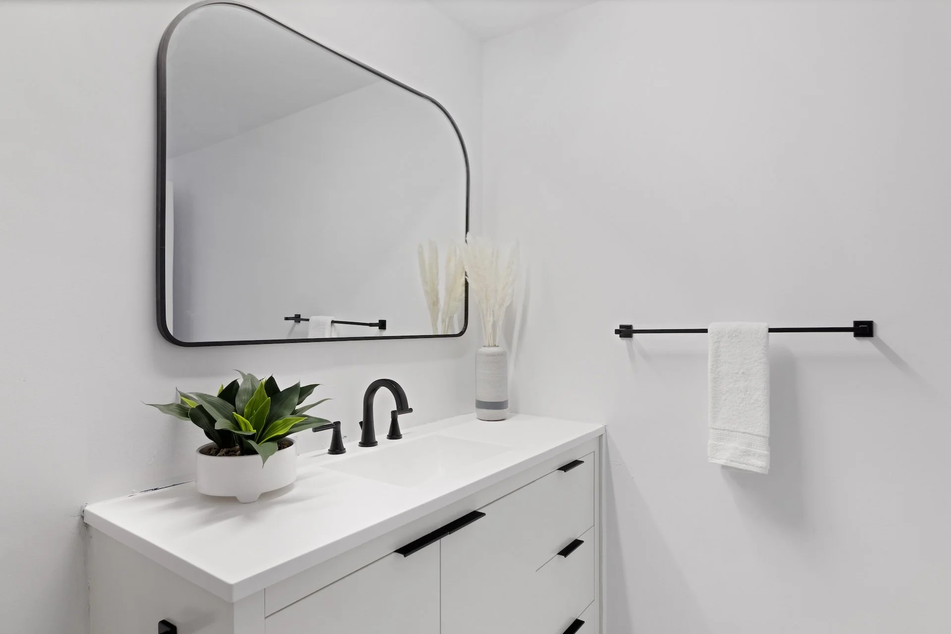 Minimalist white bathroom vanity with black hardware, a black faucet, a mirror, a potted plant, and a vase with white decorative sticks, with a white towel on a black towel bar.
