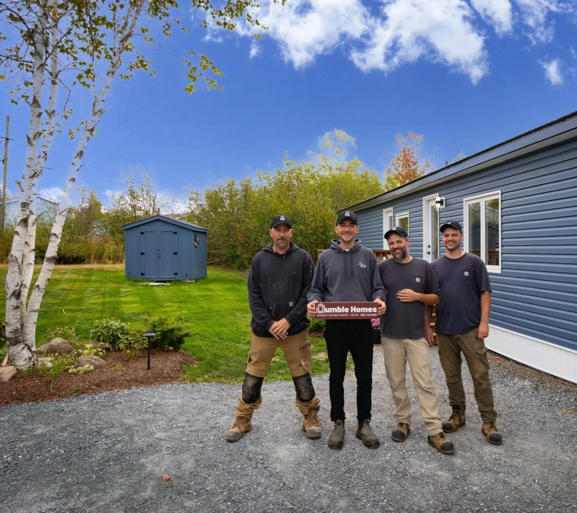 Four men standing outside in front of a blue house, with one holding a sign that says "Dumble Homes." They are smiling, wearing casual work clothes, and appear to have just completed a home construction or renovation project. The scene is set on a gr