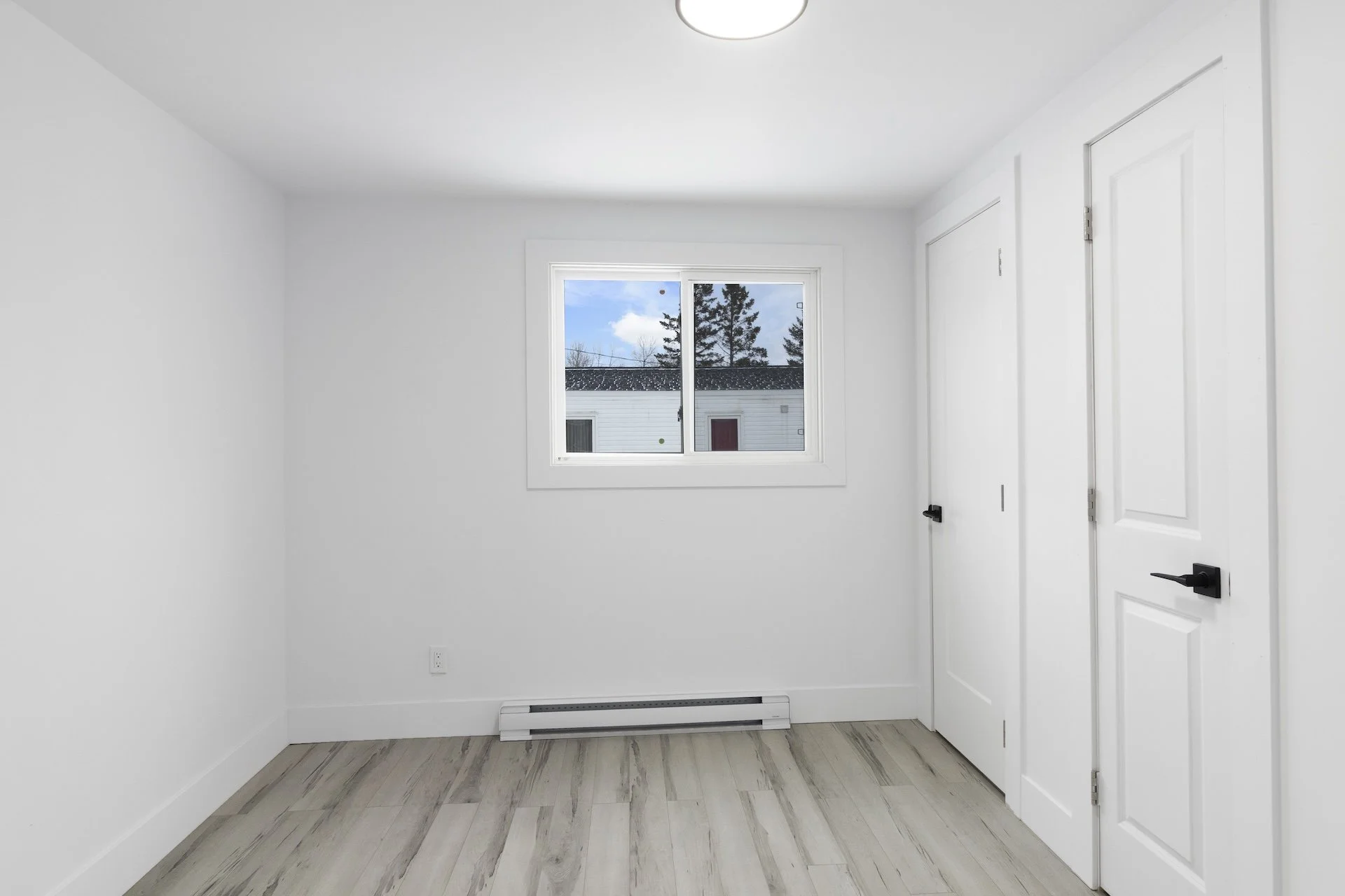 Empty white-walled room with wood flooring, a window showing trees outside, and two white closet doors with black handles.