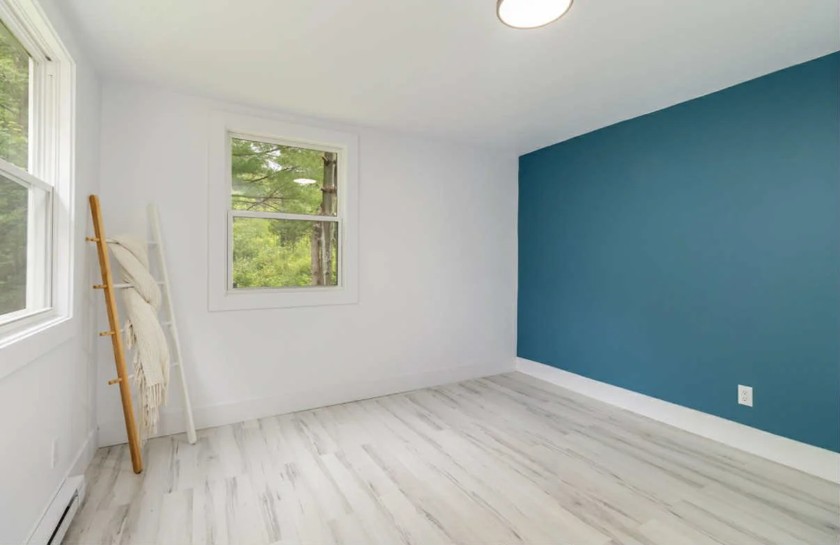 Empty room with white and teal walls, two windows showing greenery outside, and light-colored wood flooring.