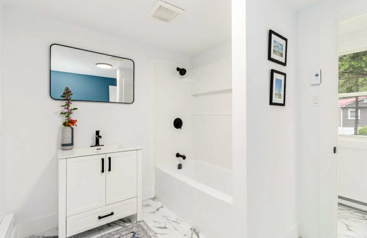 Modern bathroom with white walls, a black-framed mirror, a white cabinet with black handles, a small vase with flowers, a black faucet, a bathtub with black fixtures, and framed artwork on the wall near a window.