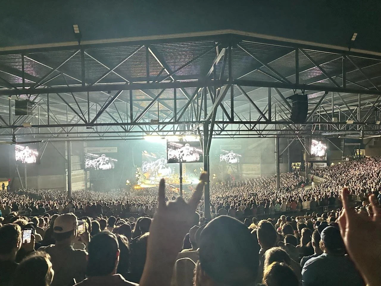 Crowd at a concert or large event in an indoor arena with stage, screens, and overhead trusses.