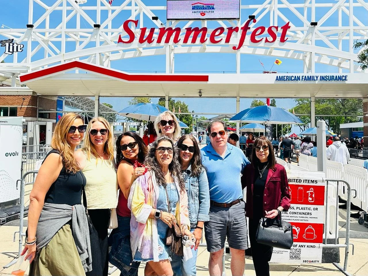 Group of seven smiling people wearing sunglasses standing outside at Summerfest, with crowds and umbrellas in the background, under a large sign that reads Summerfest.