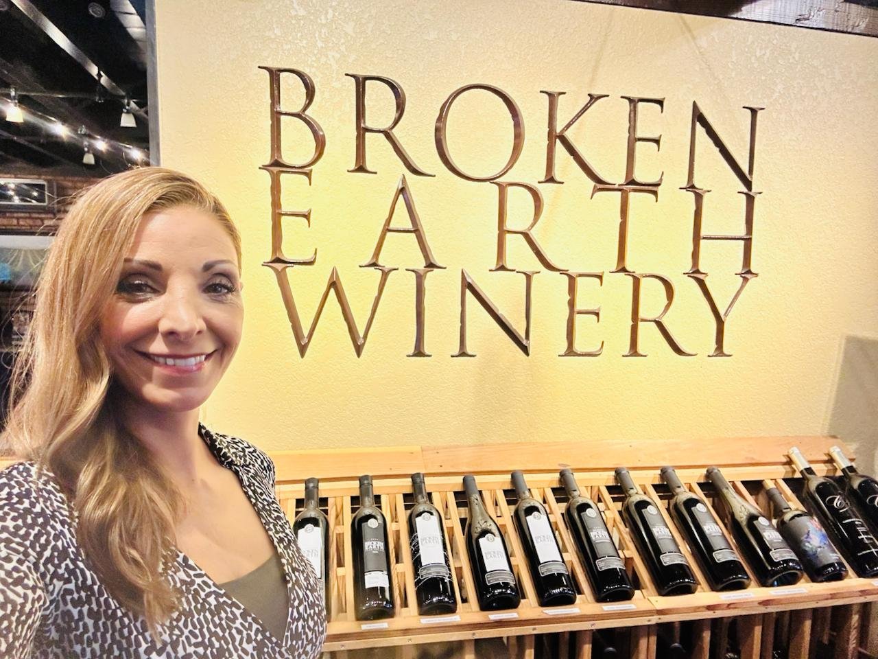 Woman with long blonde hair smiling in front of a display of wine bottles at Broken Earth Winery.