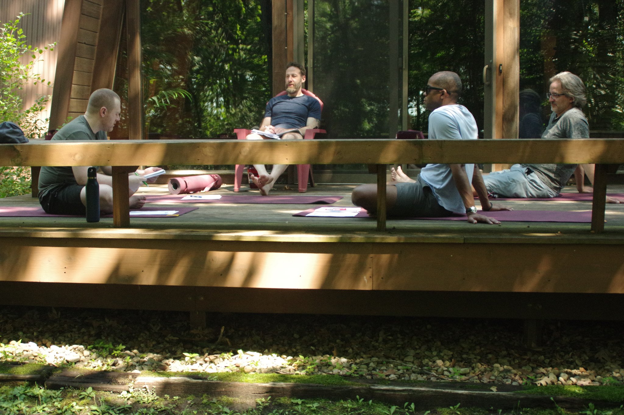 Four men are seated outdoors on a wooden deck, participating in a meditation or discussion session. They are sitting cross-legged on yoga mats with notebooks and a water bottle visible. The setting is surrounded by trees and natural greenery, with a wooden and glass structure behind them.