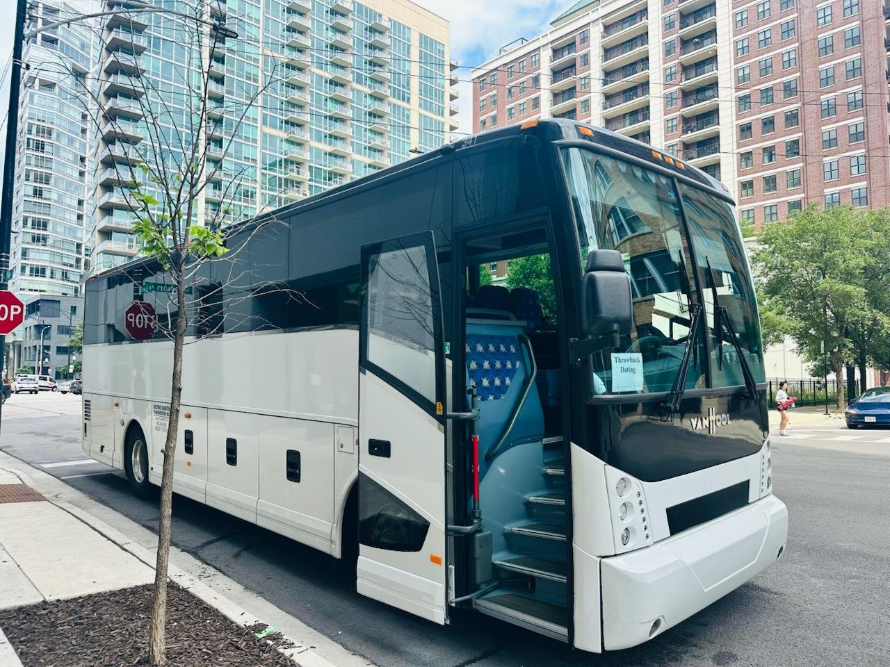 A white and black tour bus parked on city street with tall apartment buildings and trees in the background.