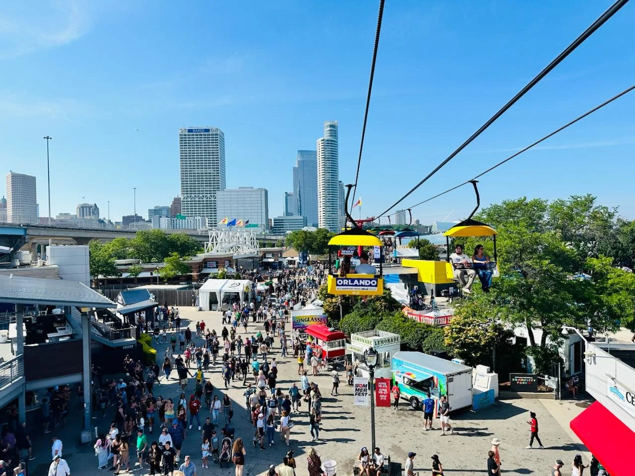 Crowd of people walking at an event with a city skyline and a cable car in the background.