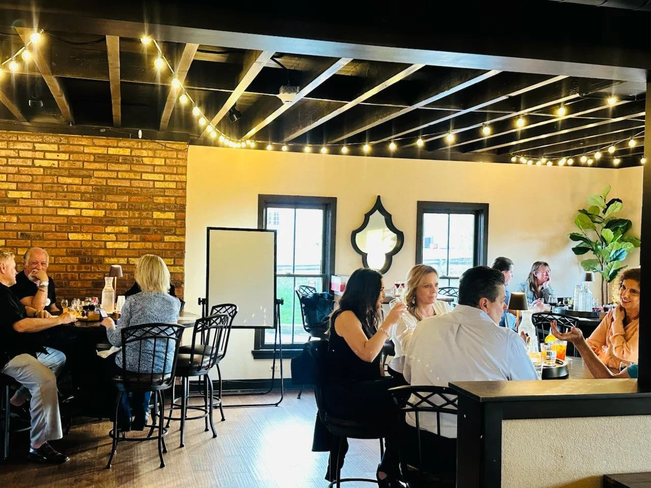 People dining in a restaurant with string lights, brick wall, and large windows.