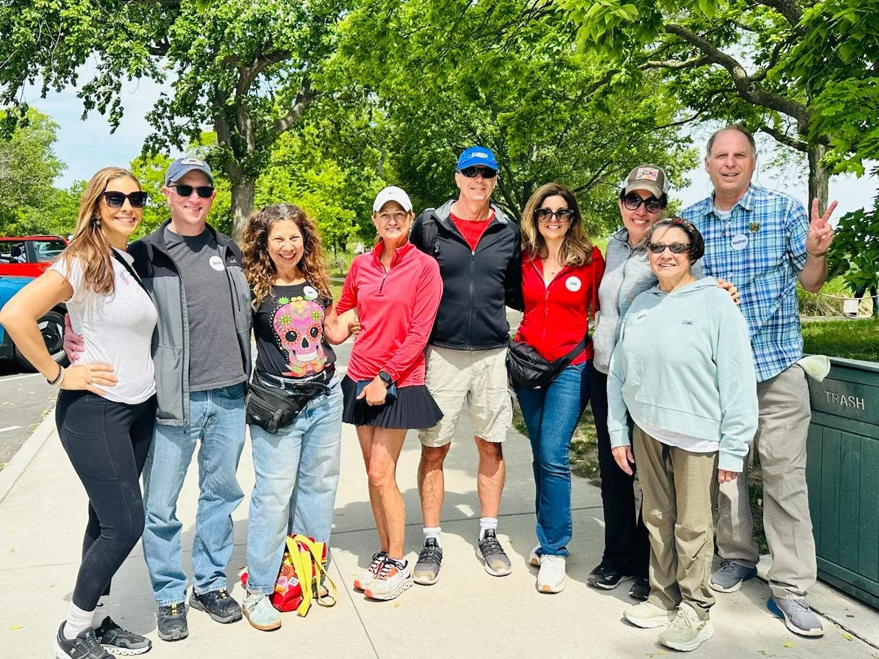 A group of ten people standing outdoors on a sidewalk, smiling and posing for a photo, with trees and a blue sky in the background — Throwback Dating singles events Chicago over 50.