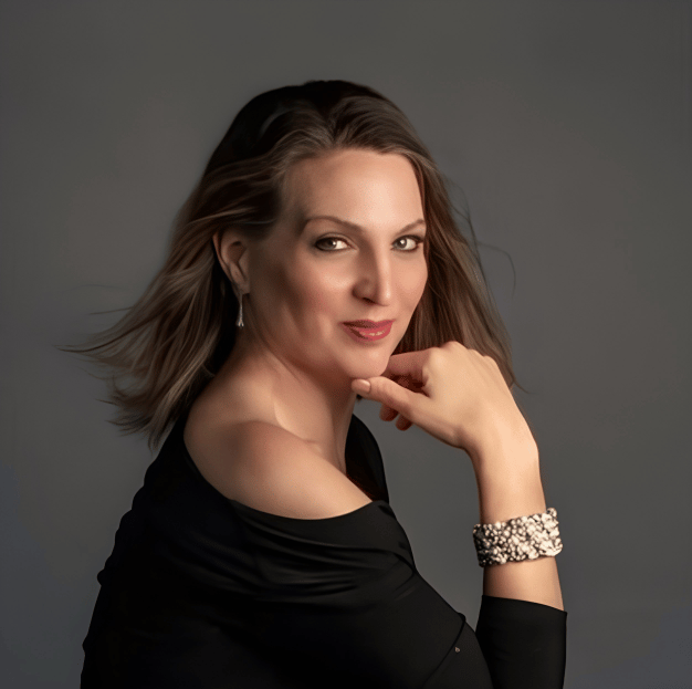 A woman with shoulder-length light brown hair and fair skin, wearing a black off-the-shoulder top, silver earrings, and a jeweled bracelet, poses with her head resting on her hand and a confident expression against a dark gray background.