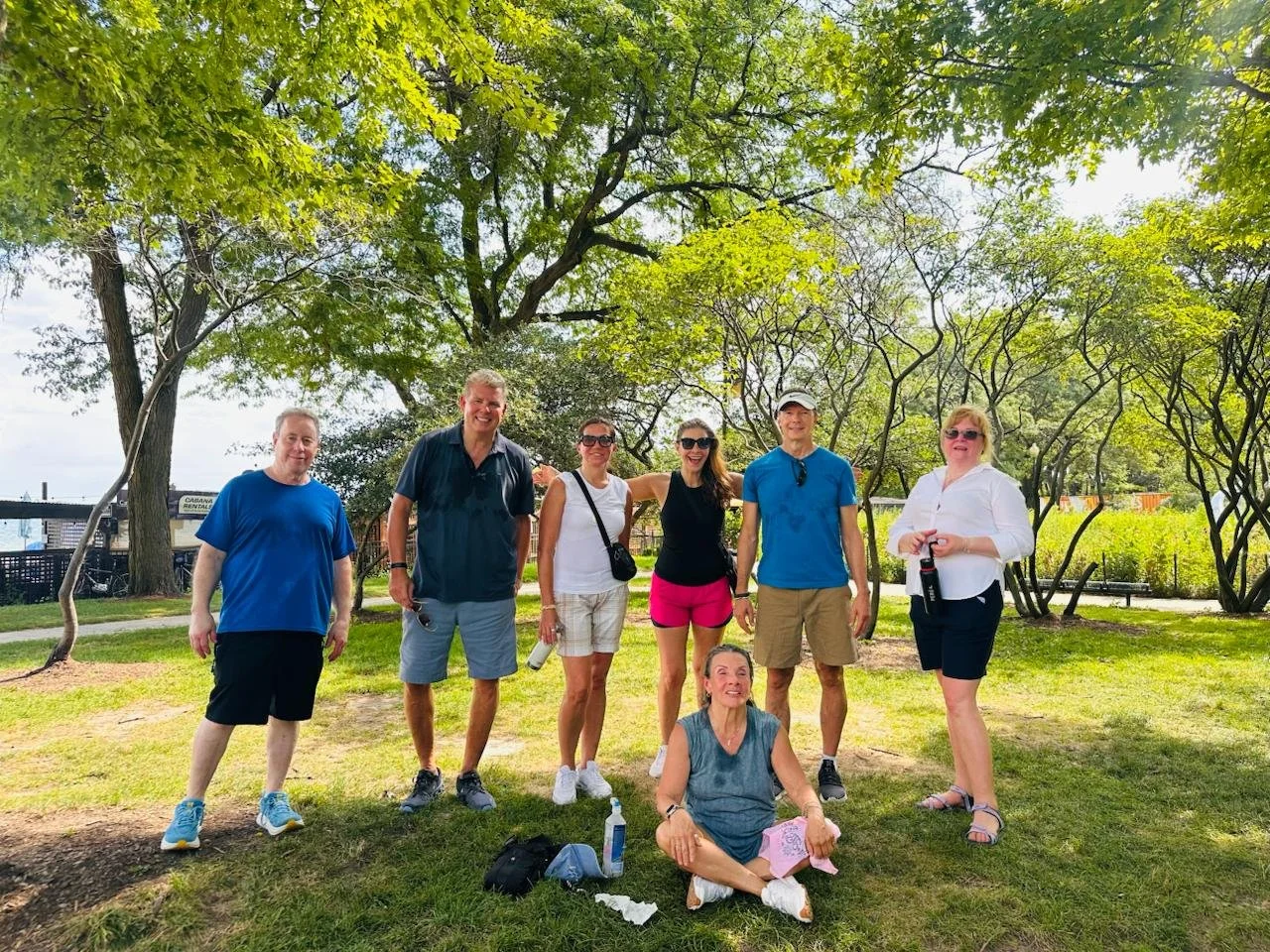 A group of seven people, six standing and one sitting on the grass, posing outdoors under green trees on a sunny day in a park.