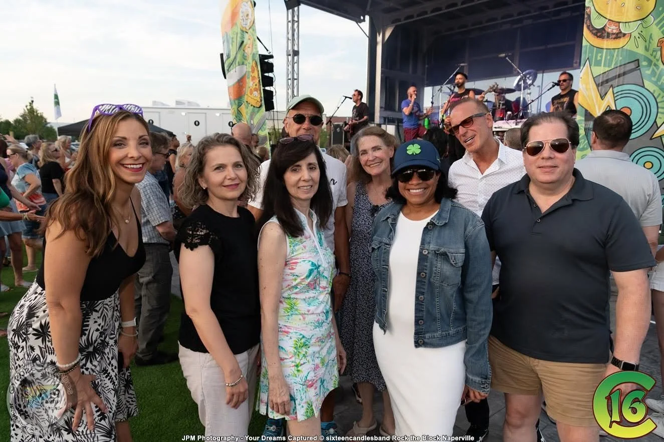 Group of people at an outdoor concert, smiling and posing for a photo, with a band performing on stage in the background.