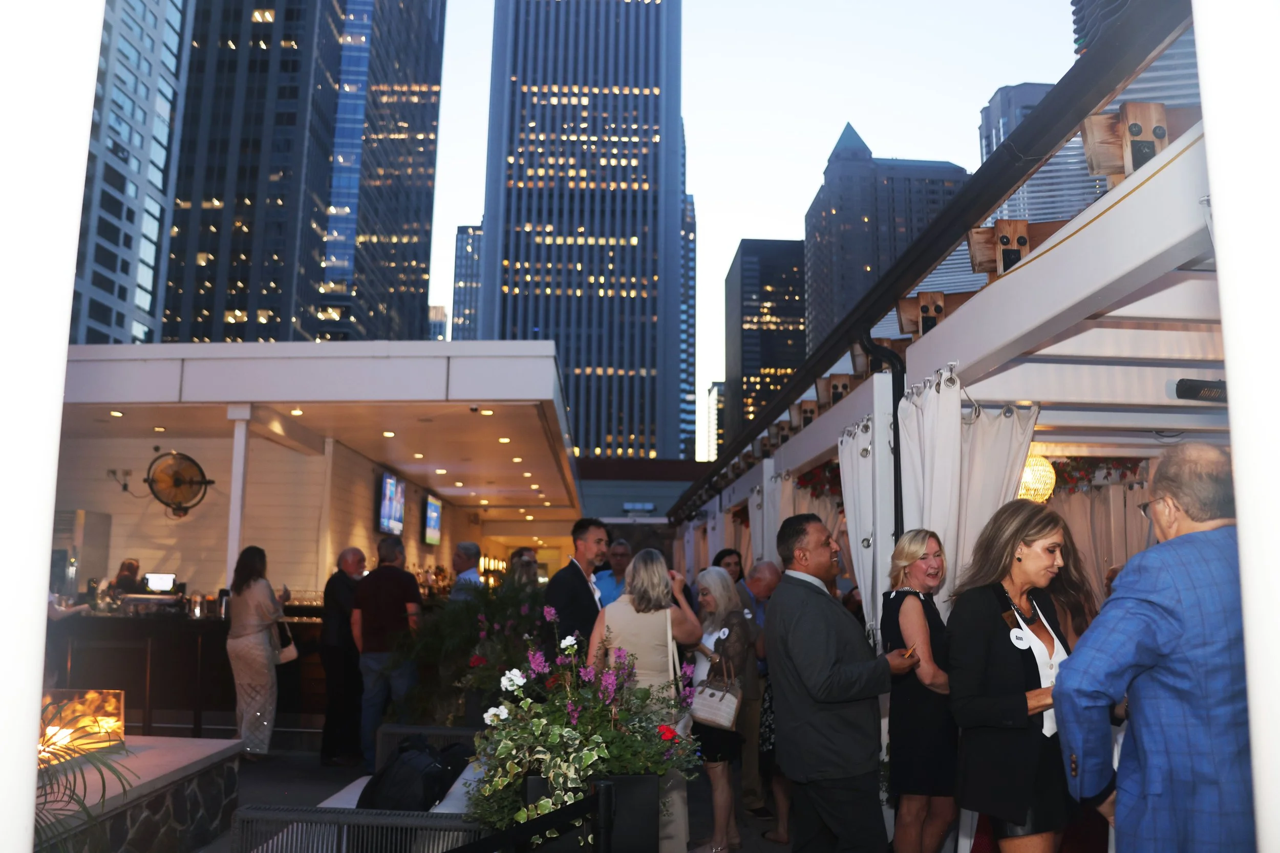 People gathered at an outdoor event in a city, with tall skyscrapers visible in the background during the evening. The scene includes a bar or service area with some attendees talking and socializing.