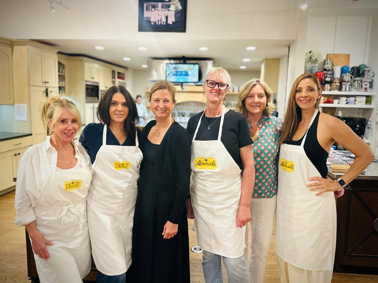 Six women standing together in a kitchen, some wearing aprons with handwritten name tags, all smiling at the camera.
