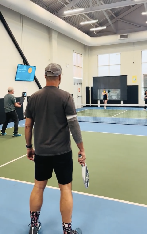 People playing pickleball indoors on a court with a net, one person preparing to serve and others watching or waiting.