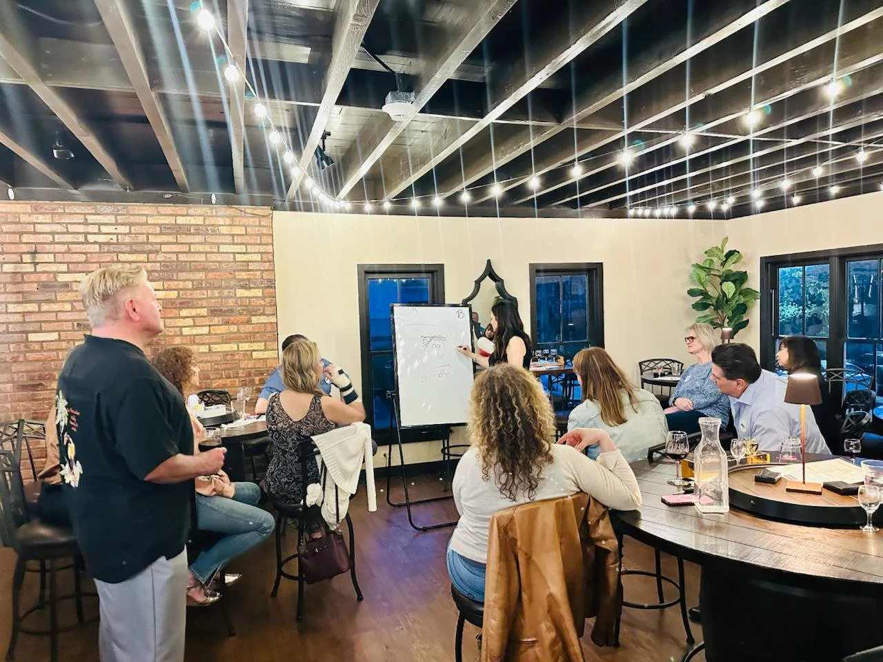 People attending a workshop or presentation in a restaurant or event space with exposed brick walls, large windows, and string lights on the ceiling.
