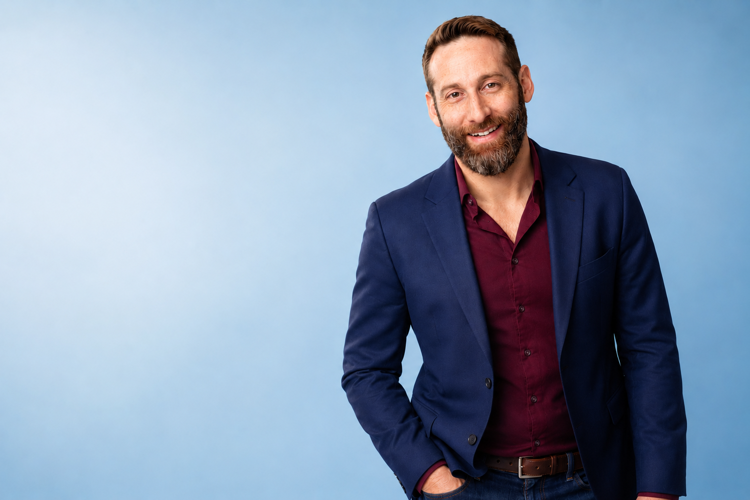 A man with a beard, wearing a navy blue blazer and maroon shirt, smiling at the camera with a blue background.