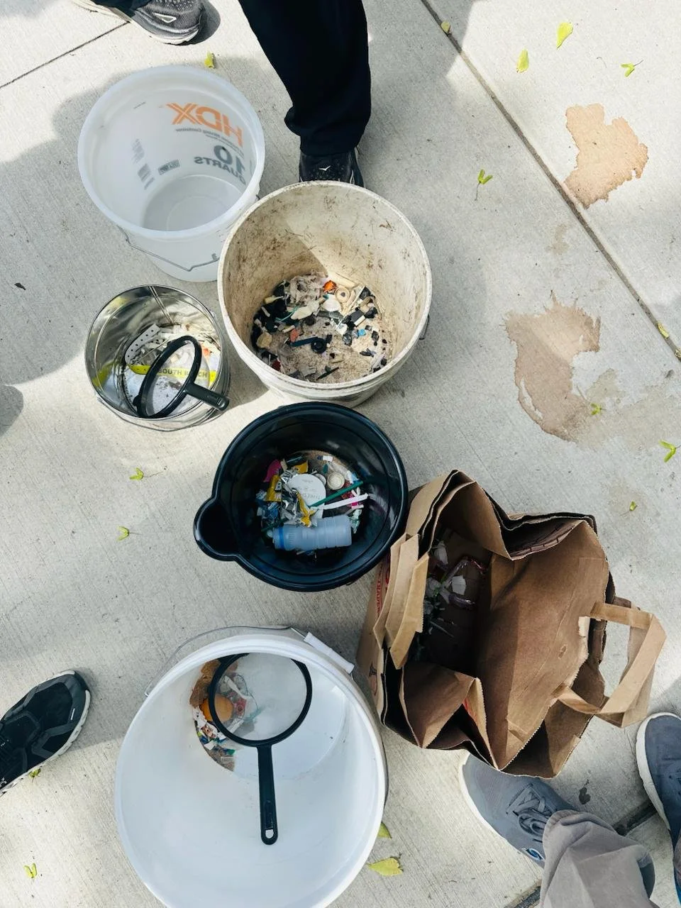 Various containers filled with collected trash, including a large white bucket, a smaller metal bucket, a black container, and a brown paper bag, with a person's feet visible nearby — Throwback Dating singles events Chicago over 50.